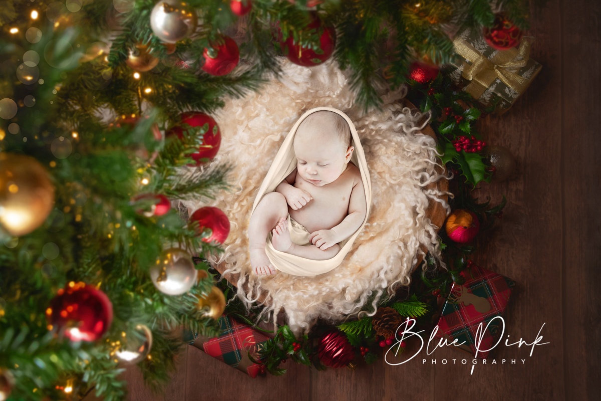 Adorable baby boy peacefully nestled on a soft white fur, gently wrapped, and surrounded by the festive glow of a Christmas tree adorned with presents.